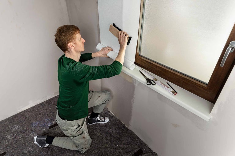 Ein Mann tapeziert eine Wand mit einer Tapetenbürste, einer Tapetenrolle, einer Schere, einem Zollstock und einem Bleistift in einem hellen Raum.