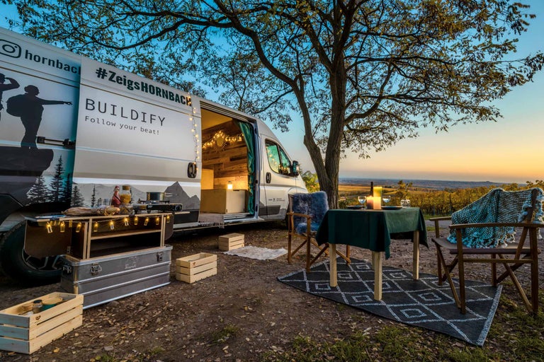 Szene mit einem umgebauten Van, einem Campingtisch und Campingstühlen im Freien.