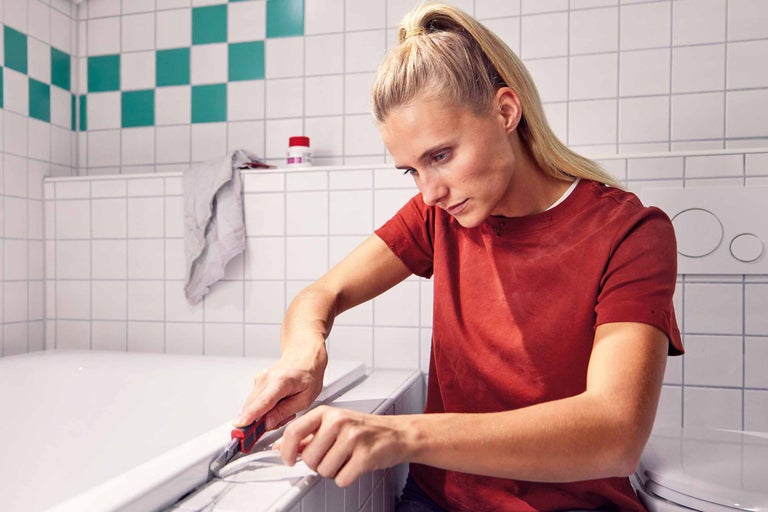 Eine Frau entfernt mit einem Cutter-Messer Silkon aus der Fuge an der Badewanne.