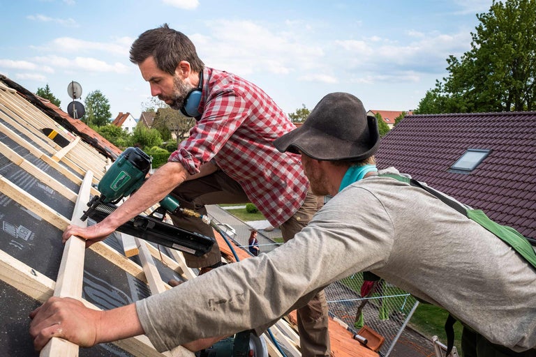 Zwei Dachdecker befestigen Dachlatten mit einem Druckluftnagler auf einem Dach.