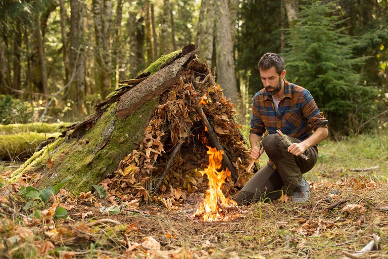 Mann entzündet ein Lagerfeuer vor einem Unterschlupf aus Ästen im Wald.