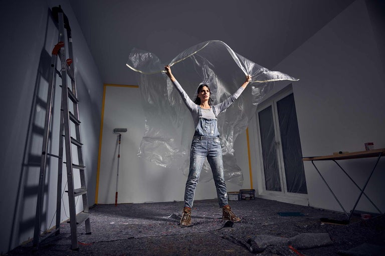 Eine Frau spannt eine Schutzfolie in einem Raum mit Abdeckvlies, Leiter und Malerutensilien.