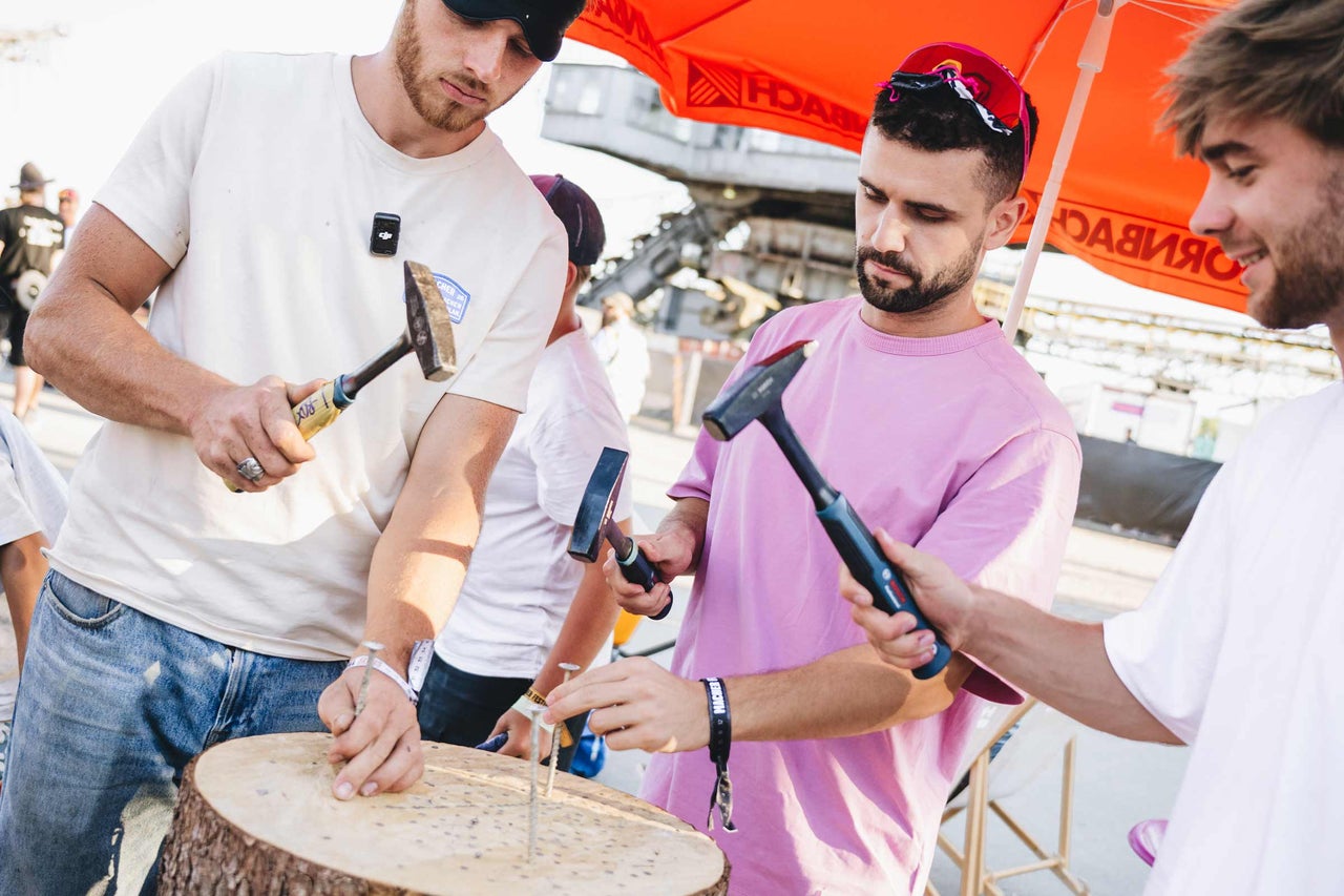Drei Männer schlagen bei einem Event unter einem Hornbach Sonnenschirm gemeinsam Nägel in einen Baumstamm.