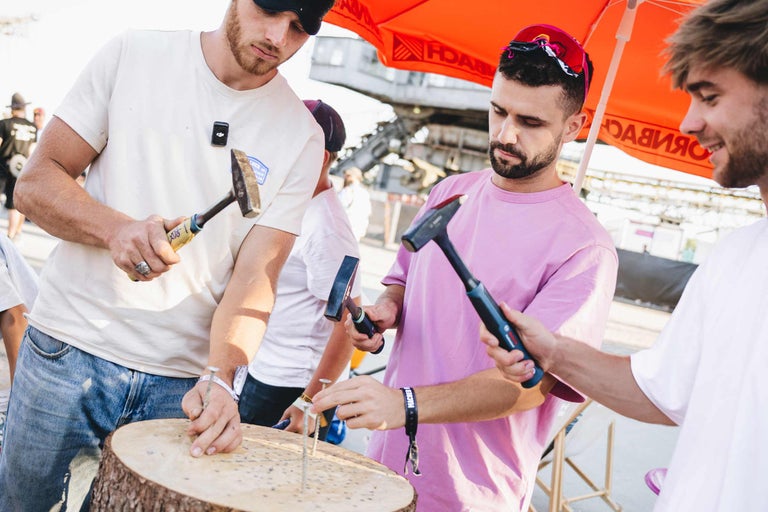 Drei Männer schlagen bei einem Event unter einem Hornbach Sonnenschirm gemeinsam Nägel in einen Baumstamm.