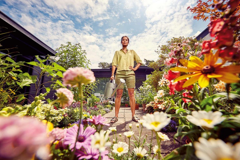 Frau mit Gießkanne steht im farbenfrohen Garten.