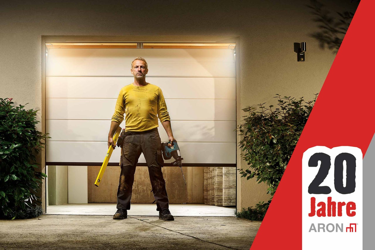 Handwerker mit Wasserwaage und Bohrmaschine vor einem ARON Sektionaltor. Siegel 20 Jahre, ARON Logo.