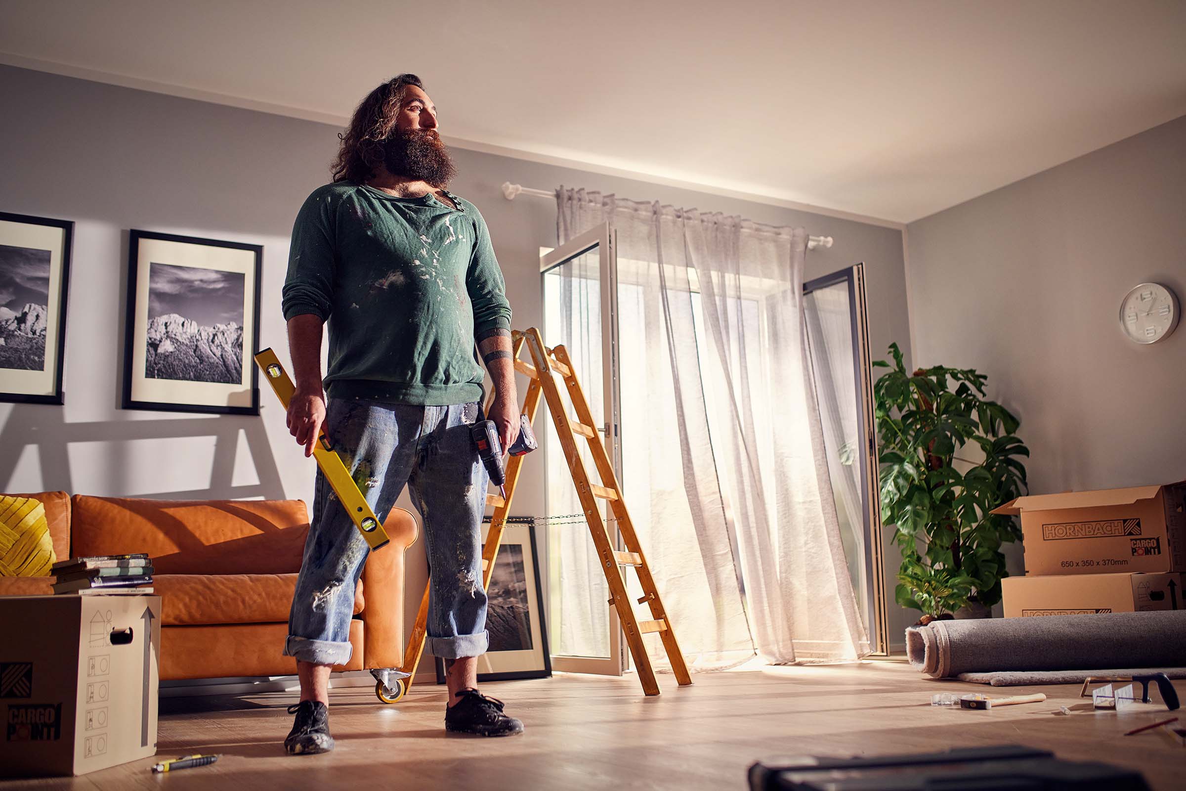 Wohnzimmer mit Mann, Wasserwaage, Leiter, Umzugskartons und Werkzeugen für Renovierungsarbeiten