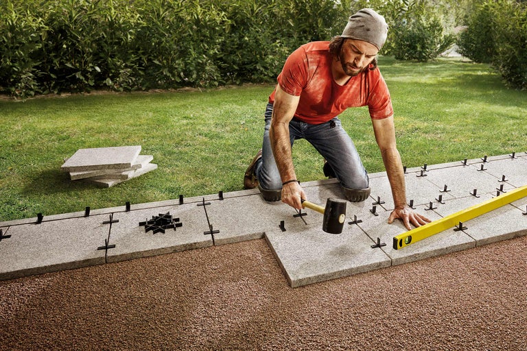 Ein Mann verlegt Steinplatten im Garten mit Hammer und Wasserwaage.