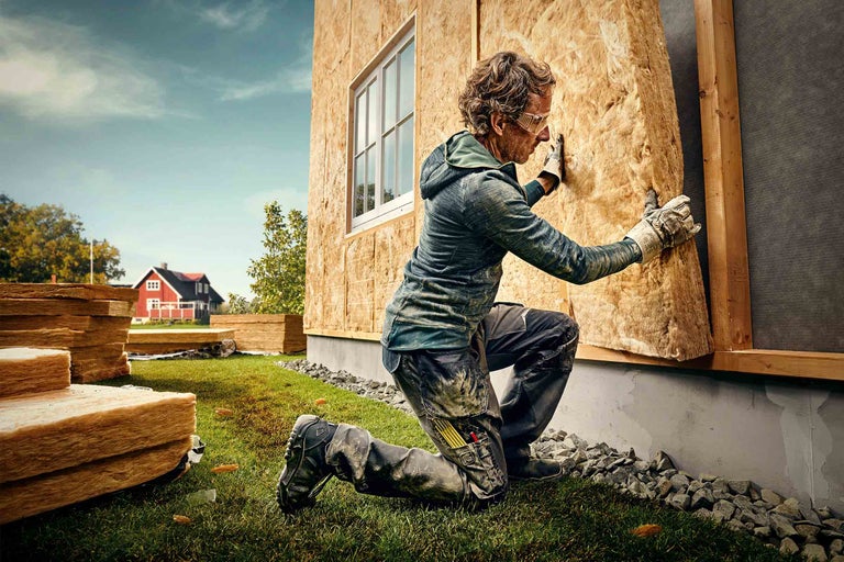 Ein Mann kniet vor einer Hauswand und befestigt Dämmwolle, wobei im Hintergrund gestapelte Dämmwolle und ein rotes Haus sichtbar sind.