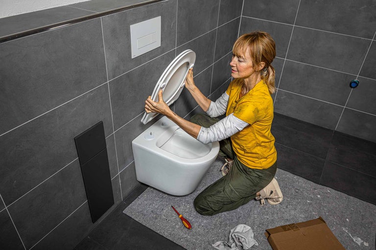 Eine Frau montiert einen WC-Sitz in einem Badezimmer mit grauen Fliesen und einem wandhängenden WC.