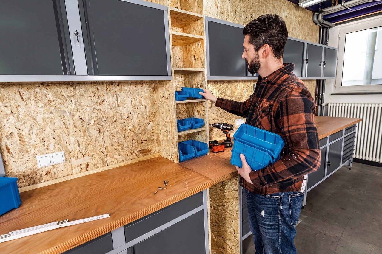 Mann organisiert Werkstatt mit blauen Sichtlagerkästen in einem OSB-Regal über einer Holz-Werkbank mit grauen Schränken.