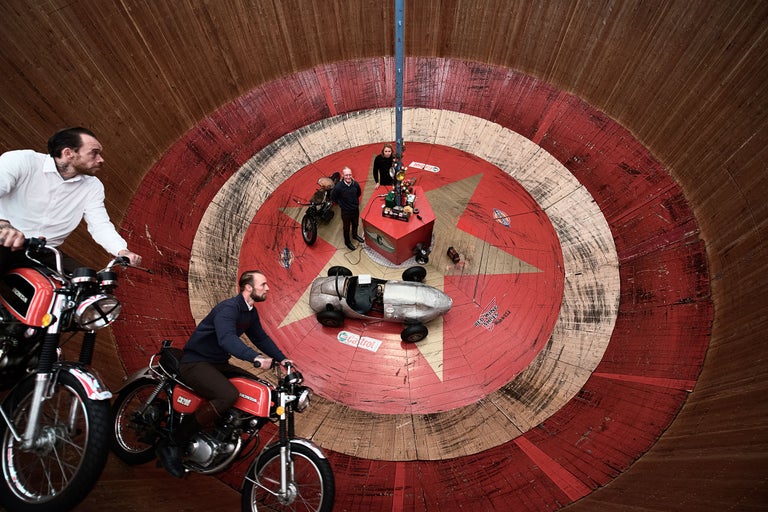 Zwei Männer fahren Motorräder in einer Steilwand. In der Mitte der Steilwand befindet sich ein Podest mit zwei Personen und einem Oldtimer.