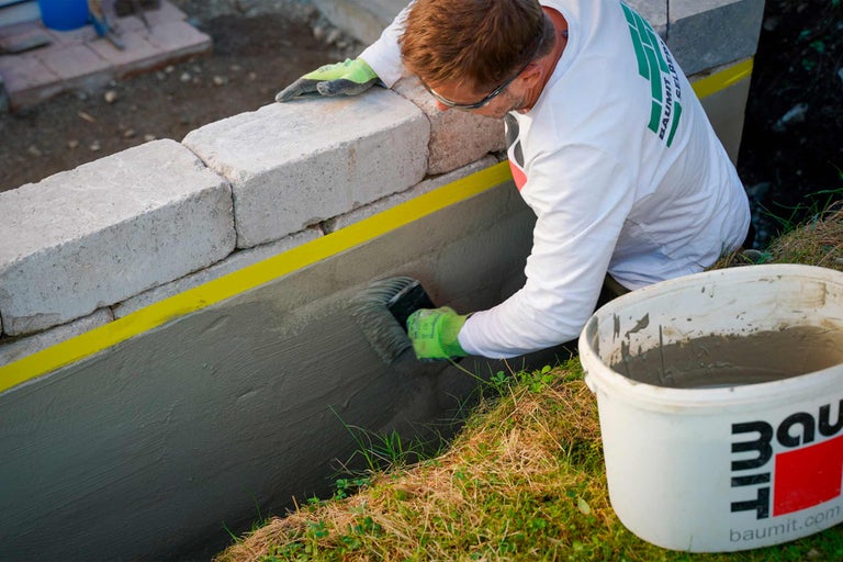 Handwerker trägt Dichtungsschlämme auf eine Steinmauer auf; Baumit Logo