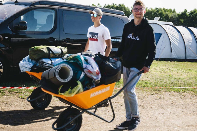 Zwei junge Männer schieben eine vollbeladene Hornbach Schubkarre mit Campingausrüstung über einen Festivalplatz.