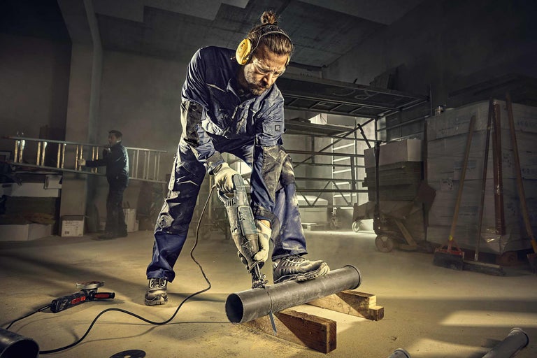 Handwerker mit Arbeitsschutz schneidet Rohr mit einer Säbelsäge. Im Hintergrund lagern Baumaterialien.