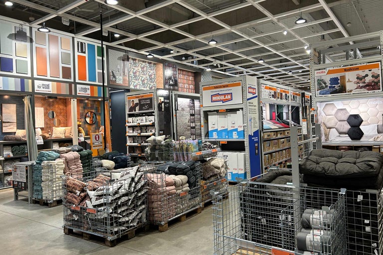 Innenansicht eines Hornbach Baumarktes mit verschiedenen Dekorationsartikeln wie Kissen, Tapeten und Paneelen in Regalen und Gitterwagen