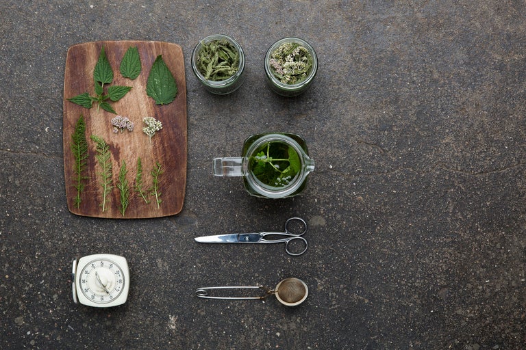 Arrangement mit Brennnesseln, Schafgarbe, Holzhackbrett, Teekanne, Küchenwecker, Teesieb und Schere