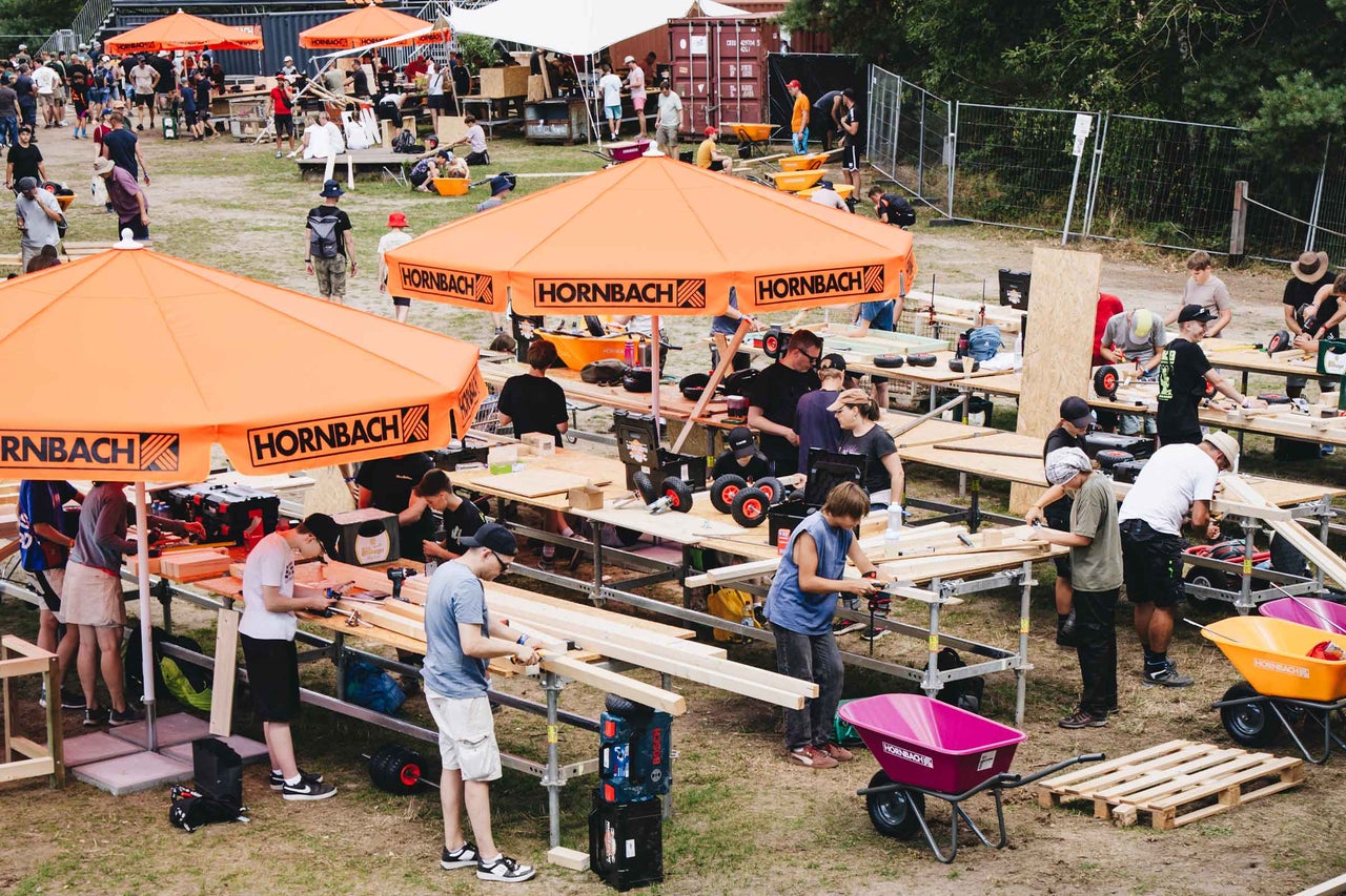 Menschen arbeiten an Werkbänken bei einem Hornbach Heimwerker Event mit Sonnenschirmen und Schubkarren im Freien.