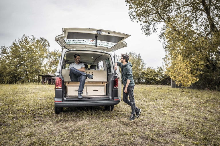 Zwei Männer an einem umgebauten Van mit Holzausbau und Schubladen auf einer Wiese.