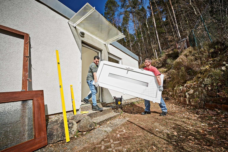 Zwei Personen tragen eine Tür vor einem Haus.