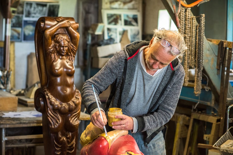 Ein Künstler bemalt eine Skulptur in einer Werkstatt. Im Hintergrund steht eine Holzskulptur einer Frau.