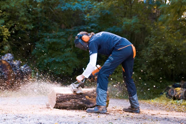Eine Person sägt mit einer Kettensäge einen Baumstamm, wobei sie Schutzkleidung und Gehörschutz trägt.