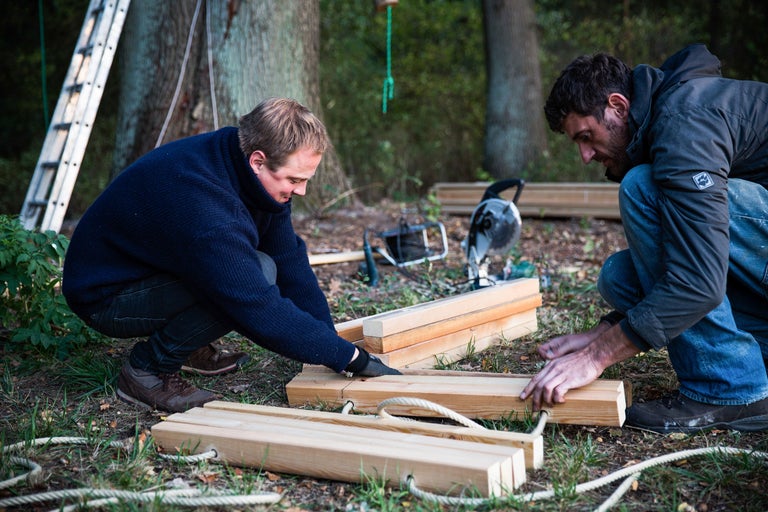 Zwei Männer arbeiten mit Holz und Seil im Freien, umgeben von Werkzeugen und einer Leiter.