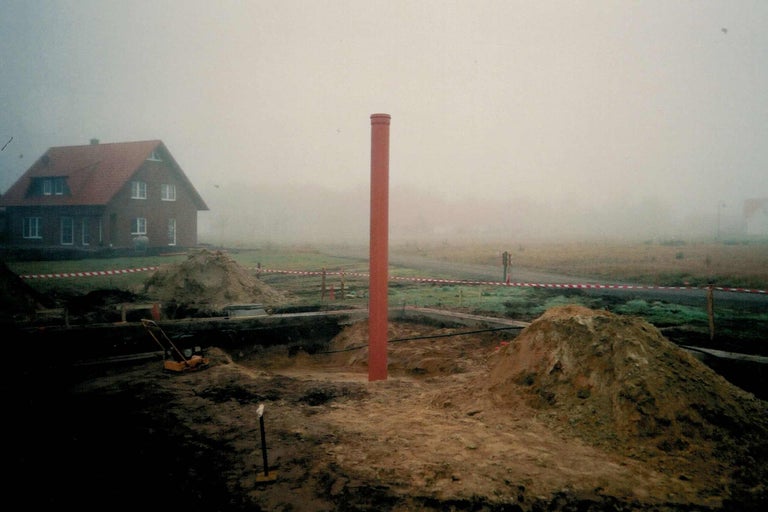 Baustelle mit Sandhaufen und einem roten Rohr im Vordergrund, Haus im Hintergrund
