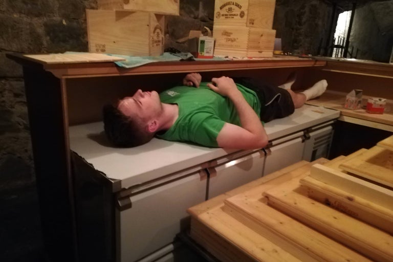 Ein Mann liegt auf einer Werkbank, umgeben von Holzkisten und Werkzeugen, in einer Werkstatt oder einem Keller.