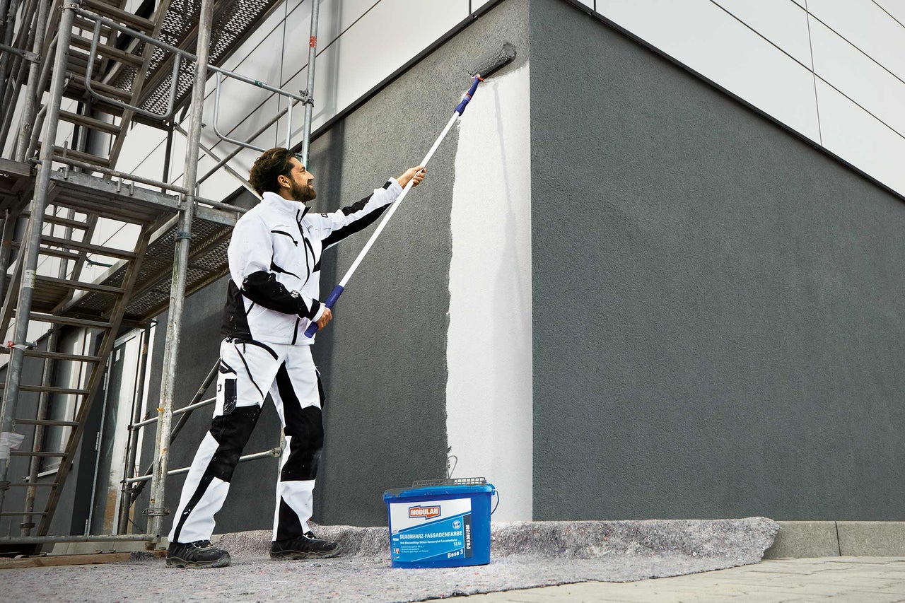 Ein Mann streicht eine Fassade mit einer Farbrolle und Fassadenfarbe. Im Vordergrund steht ein Farbbehälter der Marke Modulan.