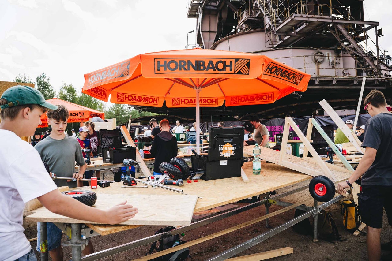 Gruppe von Jugendlichen baut Seifenkisten in einer Hornbach Werkstatt unter einem großen Sonnenschirm mit Hornbach Logo