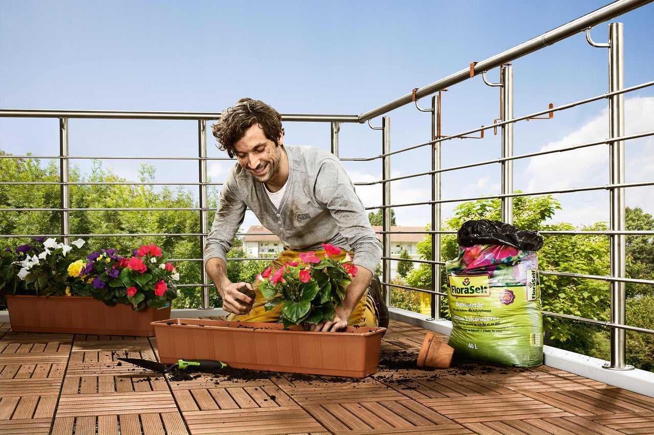 Mann bepflanzt Blumen auf einem Balkon mit Blumenerde in einem Blumenkasten.