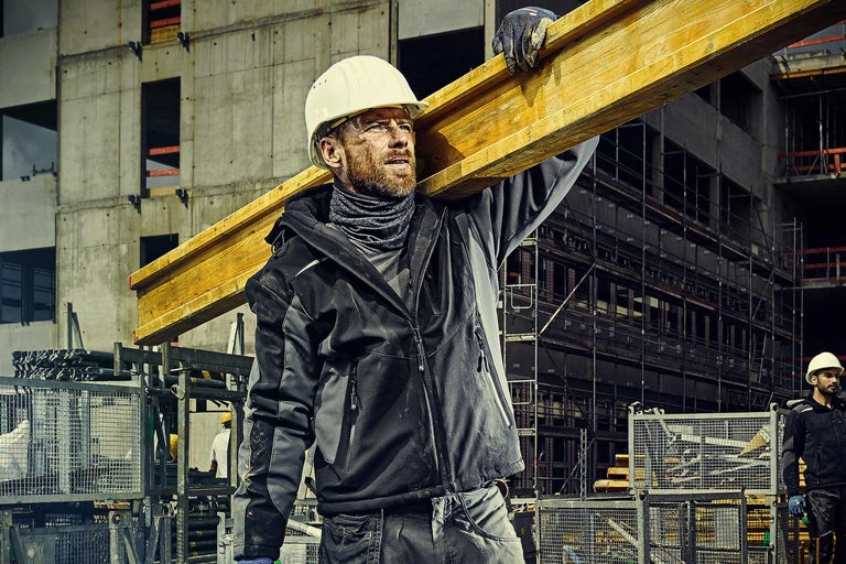 Bauarbeiter mit Schutzhelm trägt Holzbalken auf einer Baustelle