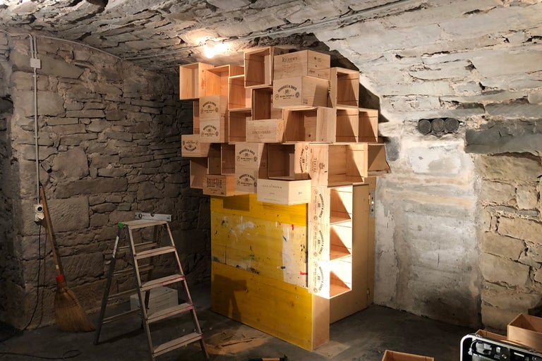 Holzkistenregal in einem Kellerraum mit Steinwänden, kombiniert mit gelben Bauelementen.