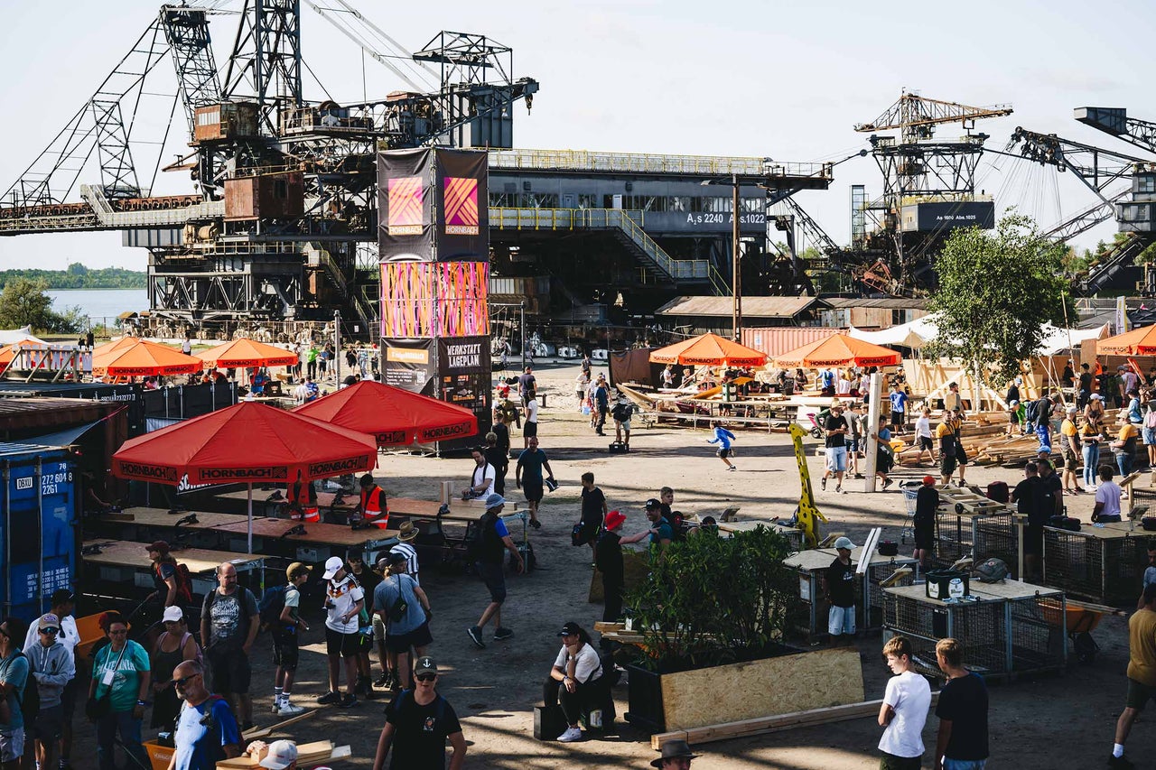 Weitwinkelansicht eines Hornbach Werkstatt Eventgeländes mit Besuchern, Werkbänken und großen Industrie Kränen im Hintergrund.