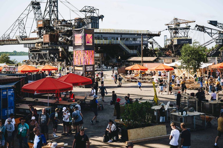 Weitwinkelansicht eines Hornbach Werkstatt Eventgeländes mit Besuchern, Werkbänken und großen Industrie Kränen im Hintergrund.