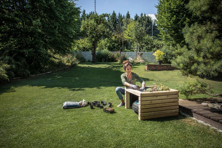Eine Frau bepflanzt einen Hochbeetkasten im Garten, umgeben von Gartengeräten und Rasen.
