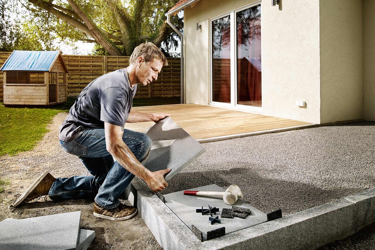 Mann verlegt Terrassenplatten im Garten mit Fugenkreuzen und Gummihammer.