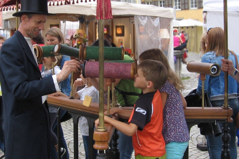 Mann mit Zylinder und Kinder benutzen Teleskope auf einem Markt.