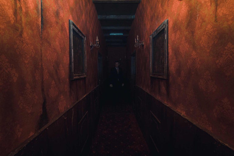 Ein dunkler Korridor mit gemusterten Tapeten, Bildern und einer Person am Ende.