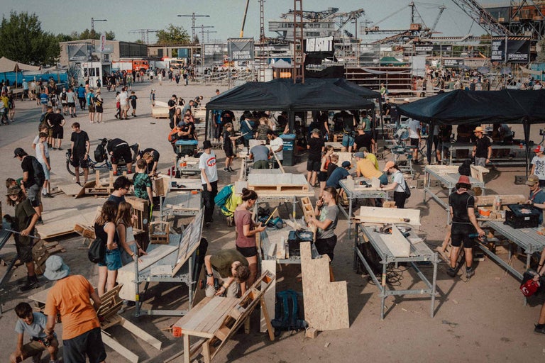 Eine lebhafte Szene zeigt Menschen, die an Werkbänken im Freien arbeiten, umgeben von einer Vielzahl von Baumaterialien und Werkzeugen vor einer Industriekulisse.