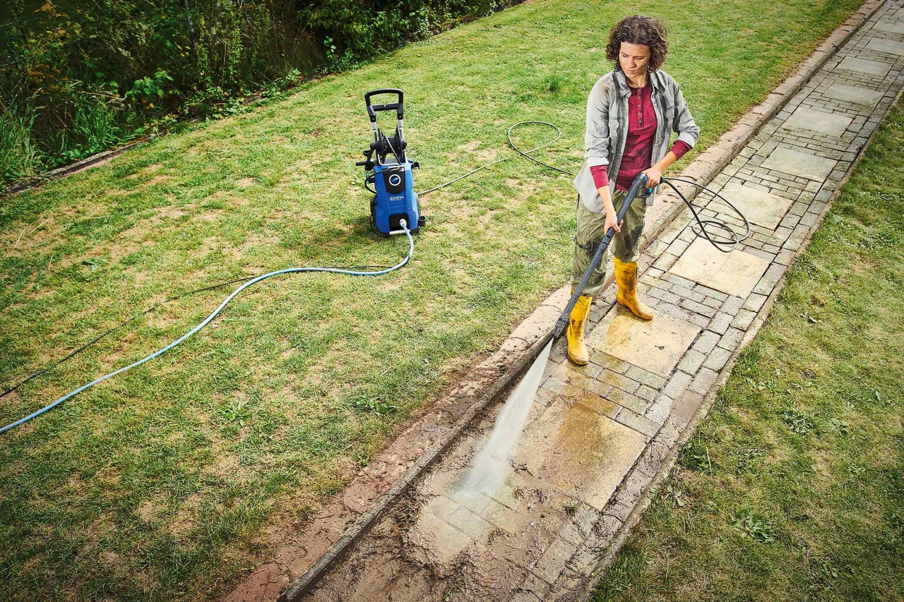 Eine Frau reinigt mit einem Hochdruckreiniger einen Gehweg in einem Garten.