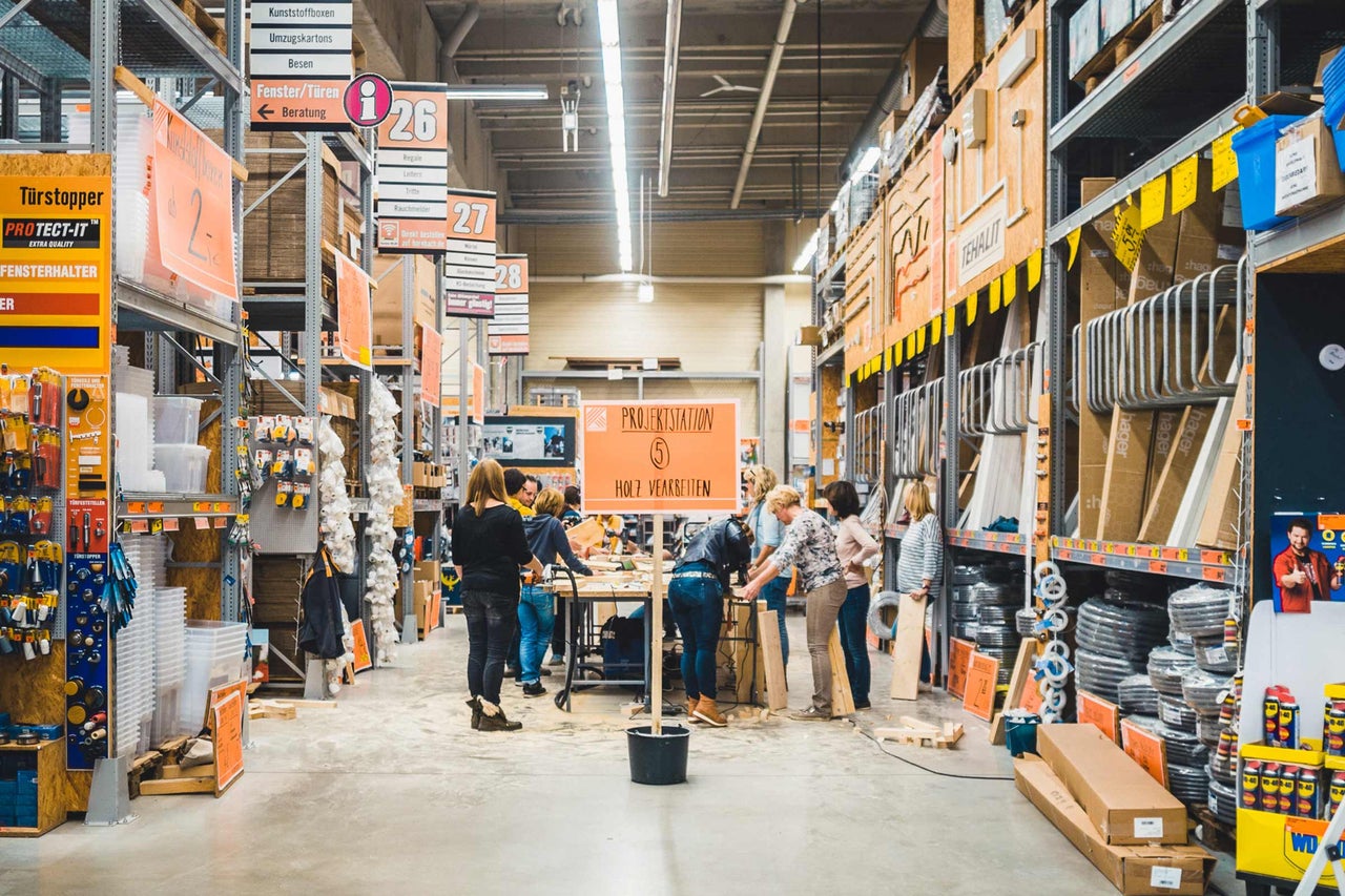 Innenansicht eines Hornbach Baumarktes mit Regalen, Waren und Kunden an einer Projektstation zur Holzbearbeitung.
