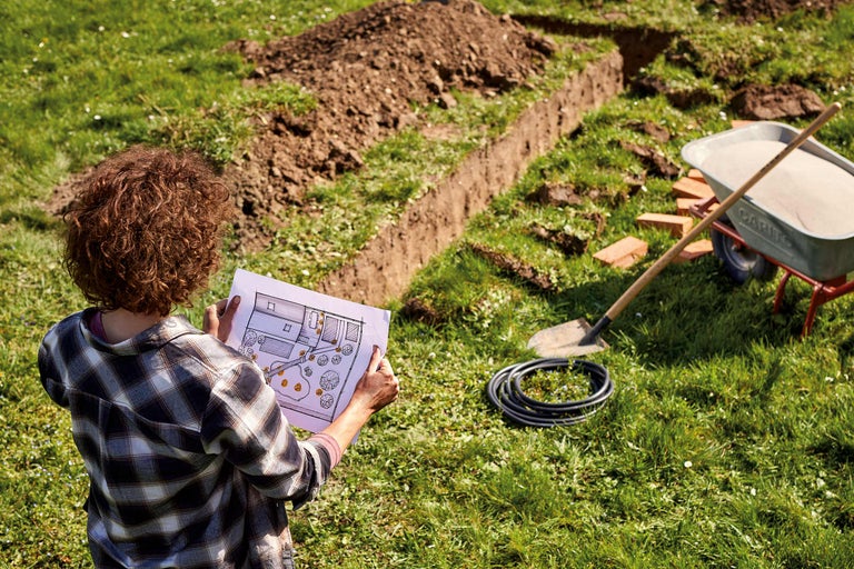 Frau betrachtet Bauplan im Garten mit Schaufel, Schubkarre, Schlauch und ausgehobenem Graben