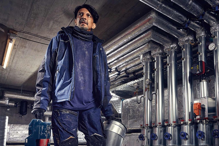 Ein Installateur mit einer Metallwanne vor Heizungsrohren in einem Kellerraum.