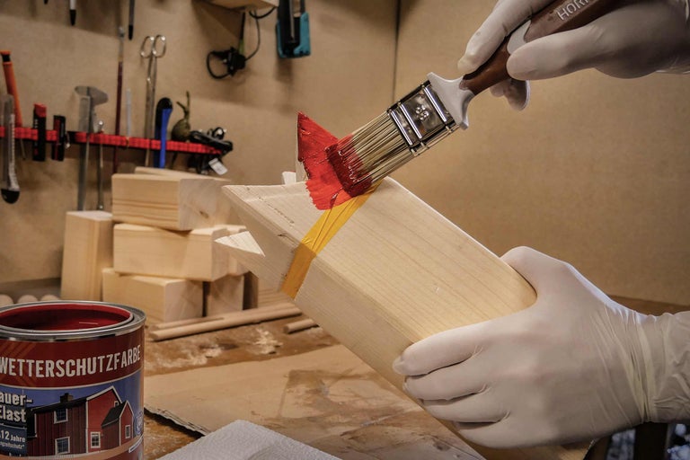 Eine Person trägt Handschuhe und streicht Holz mit einem Pinsel und roter Wetterschutzfarbe.