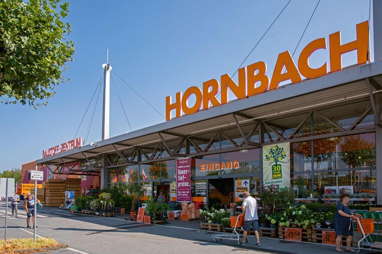 Außenansicht eines Hornbach Baumarkts mit Kunden, Pflanzen und Einkaufswagen.