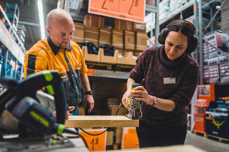 Eine Frau mit Gehörschutz sägt ein Holzbrett mit einer Stichsäge, während ein Mann zusieht.