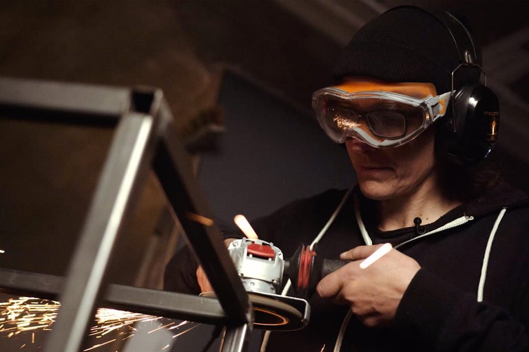 Eine Person schleift Metall mit einer Flex, während Funken sprühen. Sie trägt dabei eine Schutzbrille und Gehörschutz.