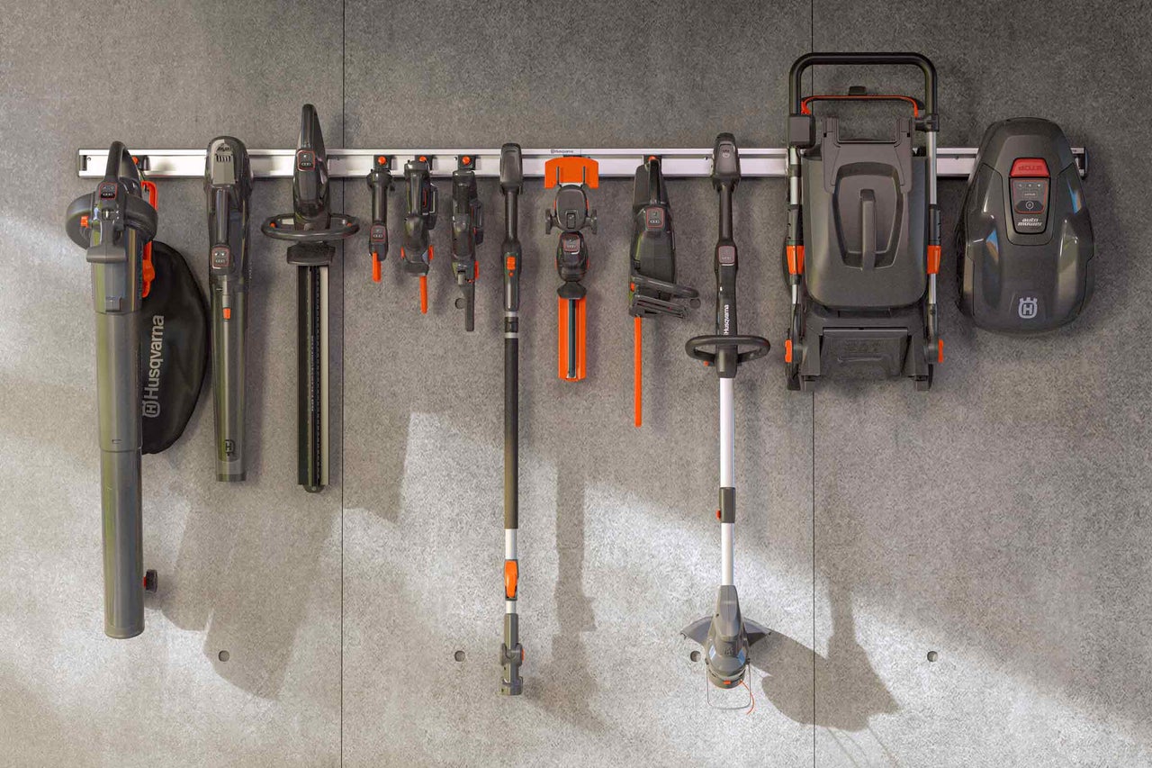 Husqvarna Gartengeräte wie Laubbläser und Rasenmäher aus Kunststoff und Metall, übersichtlich an einer Wandschiene in einer Garage organisiert.